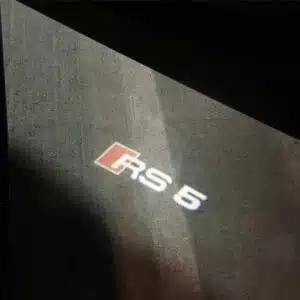High-definition LED logo projector light shining from the bottom of an Audi RS5 door onto the pavement, displaying the sharp RS5 emblem in a crisp, bright white light.